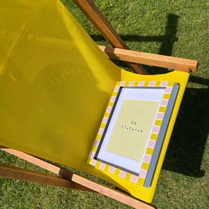 May include: A rectangular picture frame with a checkered border of alternating yellow and light pink tiles. The frame contains a pale yellow insert with the text "by florence" printed in black. The frame rests on a yellow fabric deck chair.