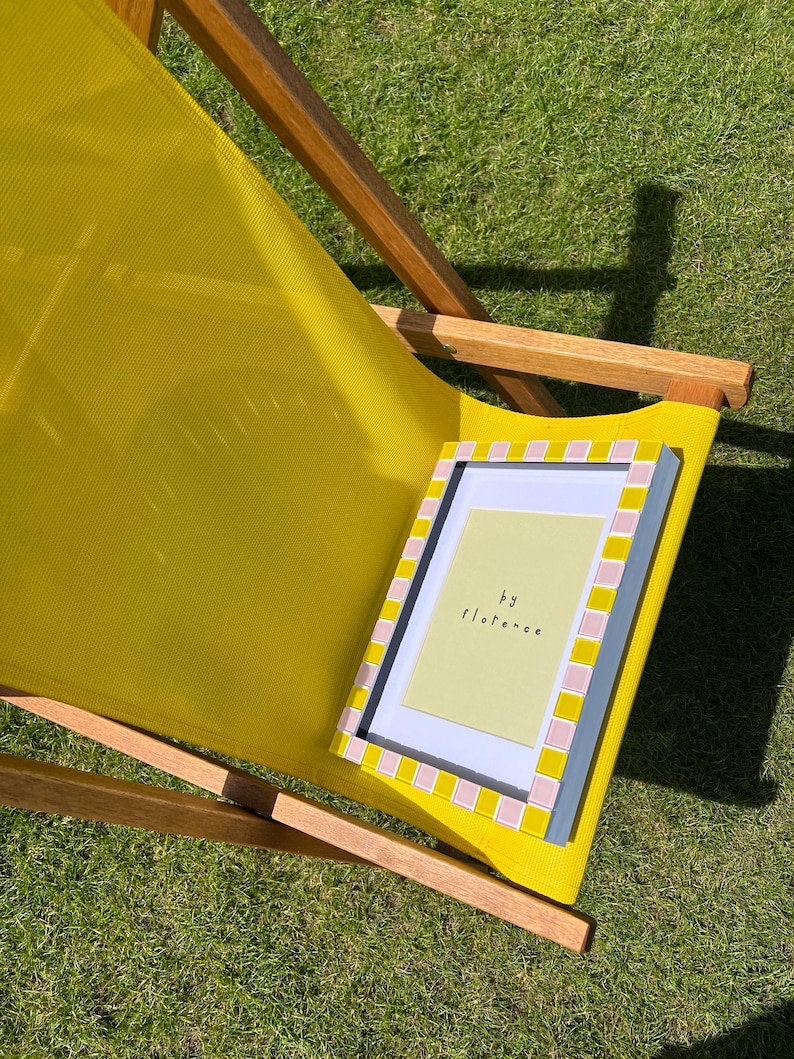 May include: A rectangular picture frame with a checkered border of alternating yellow and light pink tiles. The frame contains a pale yellow insert with the text "by florence" printed in black. The frame rests on a yellow fabric deck chair.