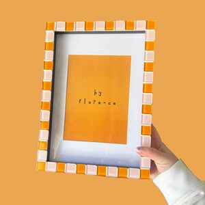 May include: A rectangular picture frame featuring a mosaic border of alternating orange and pale pink square tiles. The frame contains a white mat and a central orange print with the black text "by florence" centred in the middle of the image.