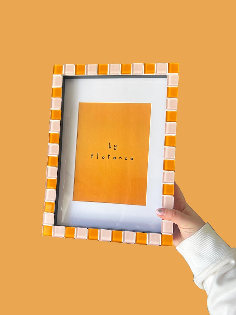 May include: A rectangular picture frame featuring a mosaic border of alternating orange and pale pink square tiles. The frame contains a white mat and a central orange print with the black text "by florence" centred in the middle of the image.