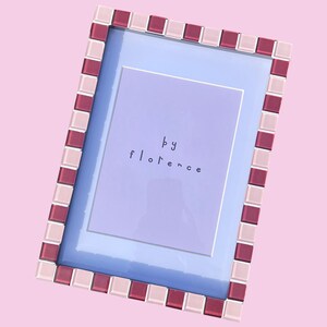 May include: A rectangular picture frame featuring a border of alternating light pink and deep magenta square glass tiles. The frame contains a light purple insert with the text "by florence" printed in the centre in a simple, dark lowercase font.