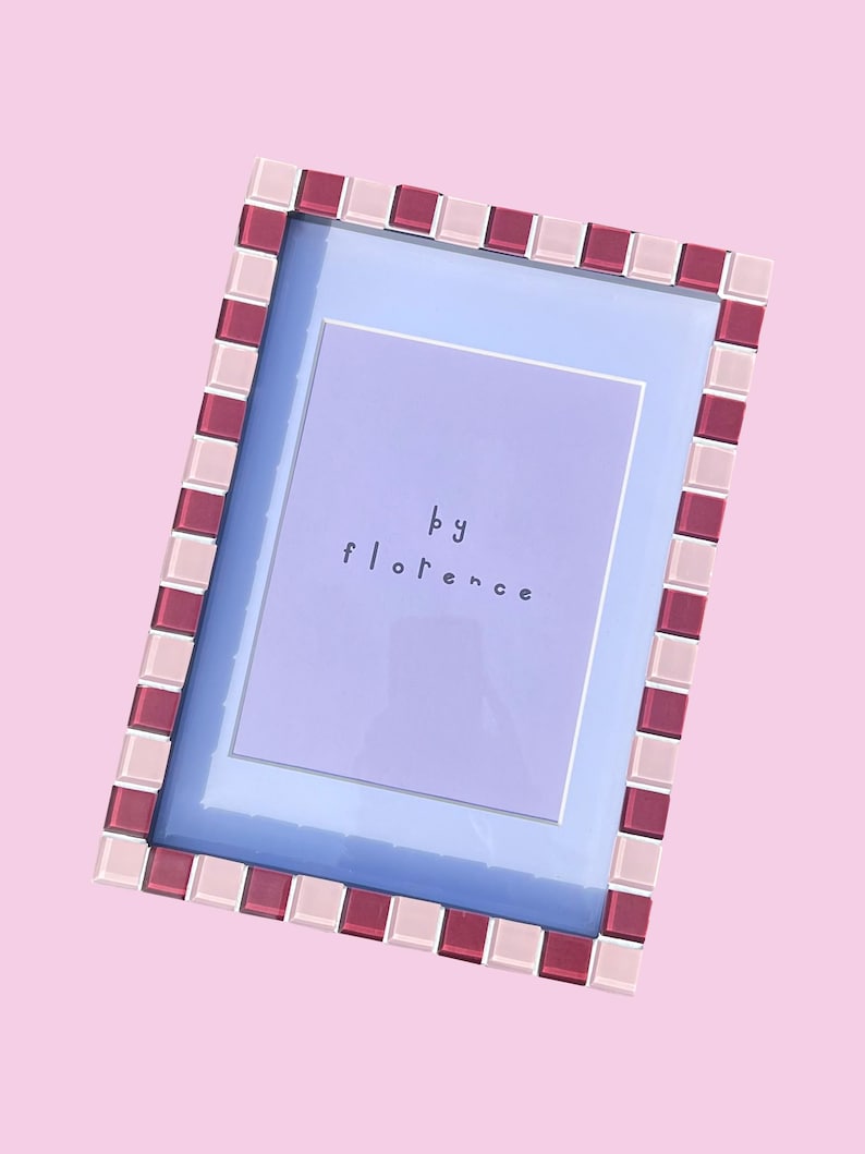 May include: A rectangular picture frame featuring a border of alternating light pink and deep magenta square glass tiles. The frame contains a light purple insert with the text "by florence" printed in the centre in a simple, dark lowercase font.
