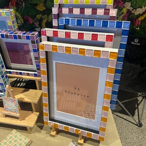 May include: A collection of rectangular picture frames featuring borders made of small, square, multi-coloured glass tiles. The frames are displayed in a tiered arrangement, with some containing inserts that read "by florence" in black text.