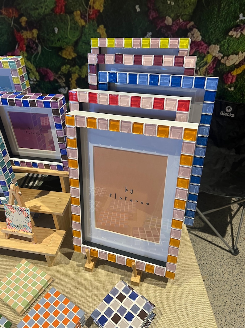 May include: A collection of rectangular picture frames featuring borders made of small, square, multi-coloured glass tiles. The frames are displayed in a tiered arrangement, with some containing inserts that read "by florence" in black text.