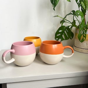 May include: Three ceramic mugs with a two-tone colour design. The mugs are in shades of pink, orange, and yellow. The mugs are sitting on a light grey table.