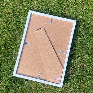 May include: A rectangular picture frame with a white border and a brown backing. The frame features a hinged support stand attached to the rear, along with four metal swivel clips used to secure the backing in place. A small pink label is visible.