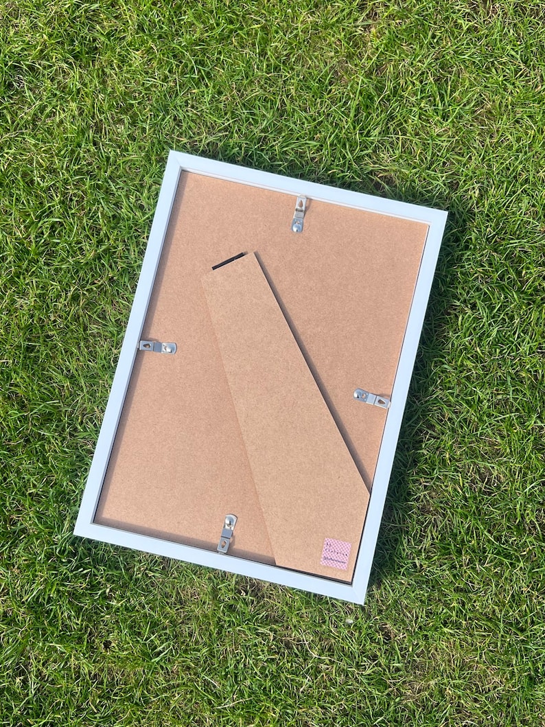 May include: A rectangular picture frame with a white border and a brown backing. The frame features a hinged support stand attached to the rear, along with four metal swivel clips used to secure the backing in place. A small pink label is visible.