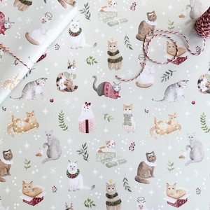 May include: Pale green wrapping paper with a repeating pattern of cats in festive outfits. The cats are shown wearing sweaters, scarves, and holiday accessories. A roll of wrapped paper and red and white twine are visible.