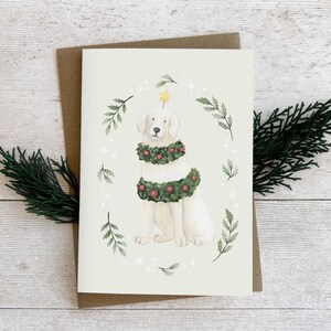 May include: A Christmas card with a watercolour illustration of a white dog wearing a festive wreath and a star. The card is set against a neutral background with sprigs of greenery.