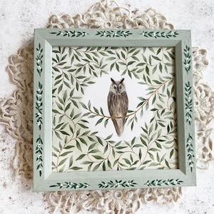 May include: A square framed wall art piece featuring a central illustration of a brown owl perched on a branch, surrounded by a dense pattern of green leaves. The light green wooden frame is decorated with a painted white leaf and berry motif.