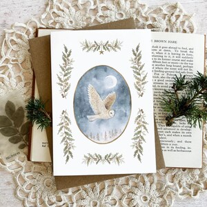 May include: A greeting card with a watercolour painting of a white owl in flight against a blue and grey background with a full moon. The card is decorated with botanical illustrations and rests on a vintage book and lace doily.