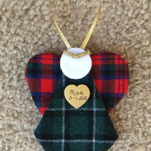 Op de afbeelding: Een handgemaakte engel ornament met rood, groen en blauw geruite vleugels. De engel heeft een witte vilten body, een gouden halo en een gouden hartvormig label met "Mom 5-1-2012". Een gouden lint is bevestigd om op te hangen.