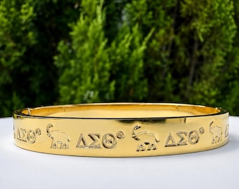 Pode incluir: Uma pulseira rígida de metal dourado com um padrão repetitivo de silhuetas de elefantes em relevo e as letras gregas Delta Sigma Theta, denotadas como ΔΣΘ, com um símbolo de marca registrada gravado na superfície externa.