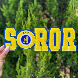 May include: A blue and yellow felt patch with the word "SOROR" in large yellow letters with blue outlines. A smaller circle with the letters "ΣΣΡ" and a poodle silhouette is centered above the word "SOROR".