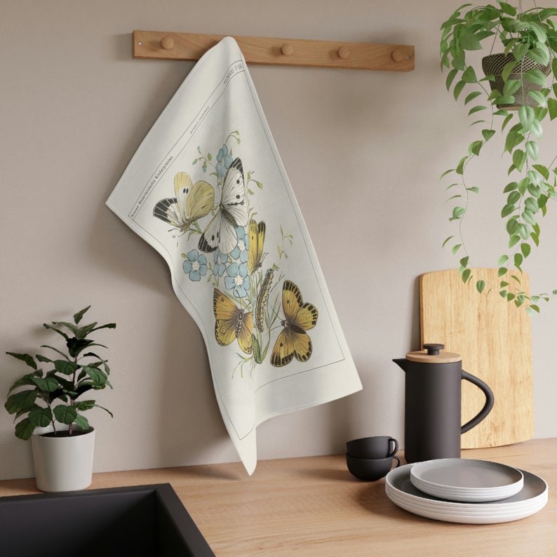 Cream colored cotton dish towel hanging on a natural wood pegged kitchen rack with printed vintage yellow butterflies, green caterpillars and flowers artwork on the front of the towel next to hanging and potted plants, coffee french press and dishes.