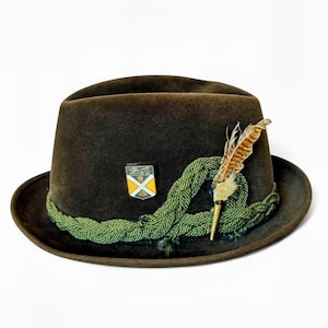May include: A brown felt hat with a green rope band and a decorative feather. The hat features a small emblem on the front and is set against a white background. A classic accessory.