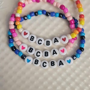 Puede incluir: Tres pulseras de cuentas con cuentas de colores. Cada pulsera tiene cuentas de letras blancas que deletrean "BCBA" con cuentas en forma de corazón. Las pulseras están sobre una superficie blanca.
