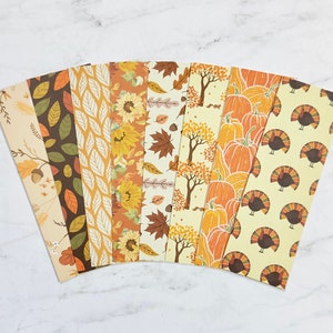 May include: A collection of autumn-themed paper sheets with various patterns. Designs include leaves, sunflowers, pumpkins, and turkeys in shades of orange, brown, and yellow. The sheets are arranged in a fan shape, showcasing the different seasonal motifs.