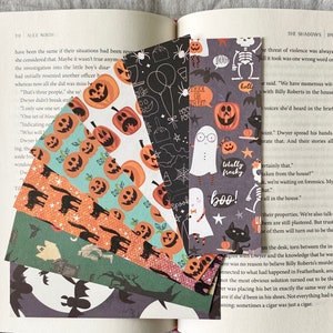May include: A collection of Halloween-themed bookmarks with various patterns. Designs include jack-o'-lanterns, ghosts, bats, and the text "Trick or Treat" and "Boo!". The bookmarks are displayed fanned out over the pages of a book.