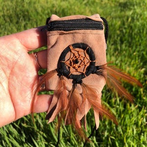 May include: A small brown suede pouch with a black zipper and a dreamcatcher design. The pouch has brown feathers attached to the bottom.