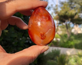 May include: A smooth, polished carnelian egg with a deep orange and red color. The egg is translucent and has a natural, organic shape.