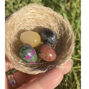 May include: A small, woven nest holds four polished gemstone eggs in shades of yellow, black, red, and green. The nest is held in a hand, with a silver ring visible. The background is blurred green grass.