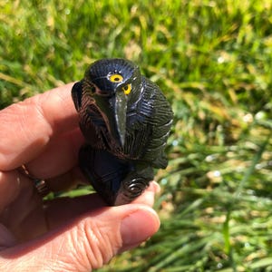May include: A black obsidian bird carving with yellow eyes. The bird is perched on a branch and has detailed carvings on its feathers.