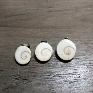 May include: Three round, off-white pendants with a spiral design, each with a silver-colored bail. The pendants are arranged in a row against a dark wood-grain surface. The spiral design is a light brown color.