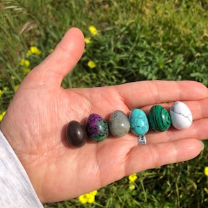 May include: A hand holding six polished gemstone eggs. The eggs are in a row from left to right: brown, green and purple, gray, turquoise, green and black, and white.