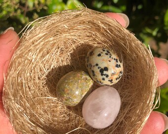May include: Three tumbled stones, a pink rose quartz, a green dalmatian jasper, and a black and white dalmatian jasper, are nestled in a small woven bird's nest.