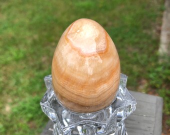 May include: A light brown, egg-shaped, polished onyx stone sits on a clear glass, star-shaped stand.