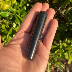 May include: A black, cylindrical stone wand with a smooth, polished surface. The wand is held in an open hand against a backdrop of green foliage, suggesting a natural or spiritual theme.