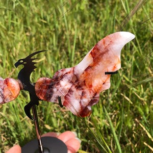 May include: A red and white crystal dragon sculpture with wings, standing on a black metal stand. The dragon is made of a natural stone with a unique pattern.