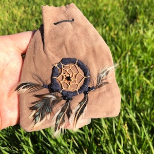 May include: A small brown suede pouch with a dreamcatcher and feathers. The dreamcatcher is made of black and gold thread and has black beads.