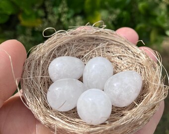 May include: A small, round, woven nest made of light brown natural fibers containing five smooth, oval-shaped, translucent white quartz stones. The stones have a polished, glossy finish and are arranged closely together within the center of the nest.
