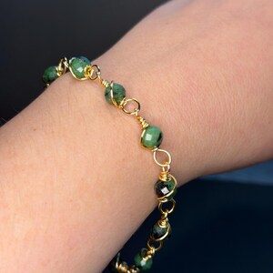 May include: A bracelet featuring faceted green and dark-speckled round beads individually wrapped in gold-toned wire. The beads are connected by small gold-toned wire loops, creating a delicate chain design that encircles the wrist.