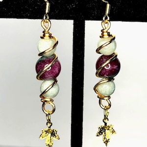 May include: A pair of dangle earrings featuring gold-toned wire wrapped around three spherical beads. The top and bottom beads are pale green, while the center bead is deep purple. A small gold-toned maple leaf charm hangs from the bottom.
