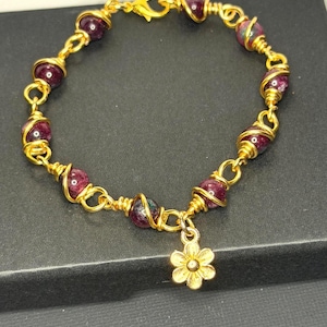 May include: A gold-toned wire-wrapped bracelet featuring round, deep magenta-colored beads interspersed with gold metal links. The bracelet includes a small, six-petaled flower charm hanging from one of the links and secures with a lobster claw clasp.