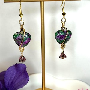 May include: A pair of dangle earrings featuring heart-shaped stones with mottled green and purple patterns. Each stone is wrapped in gold-toned wire and hangs from a gold-toned hook, with a small, faceted, light purple crystal bead dangling below.