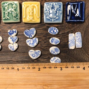 May include: A selection of ceramic tiles and shapes in various colours and designs. The tiles include letters, hearts, and oval shapes, all with blue and white patterns. The ruler shows measurements in centimetres.