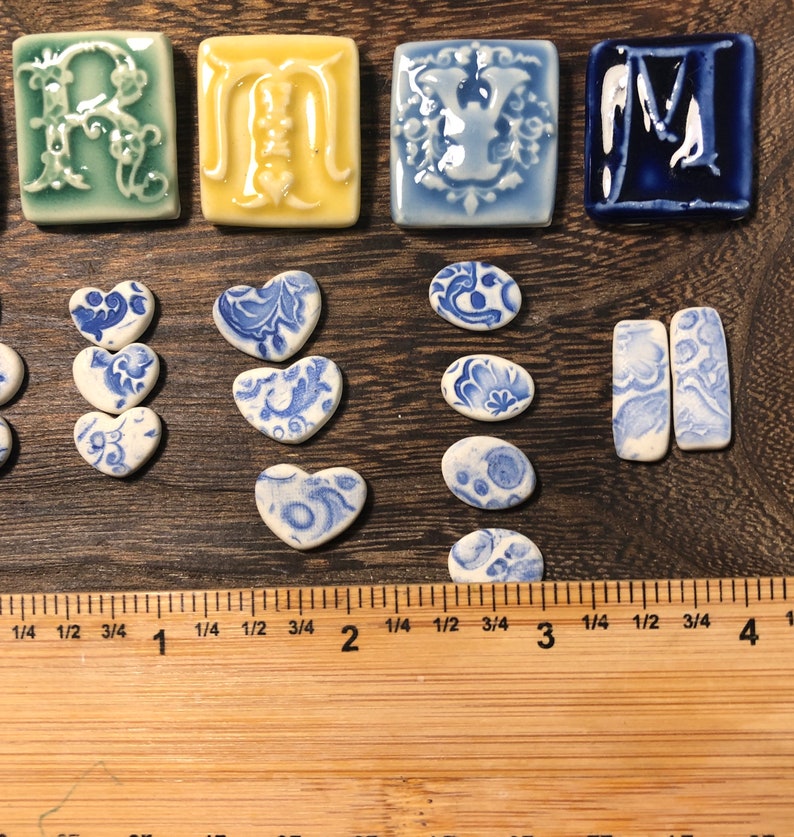 May include: A selection of ceramic tiles and shapes in various colours and designs. The tiles include letters, hearts, and oval shapes, all with blue and white patterns. The ruler shows measurements in centimetres.