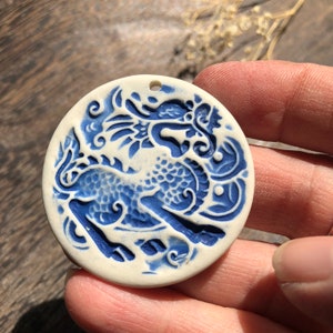Op de afbeelding: Een witte keramische hanger met een blauw en wit ontwerp van een draak. De draak is opgerold met zijn kop naar de zijkant gedraaid. De hanger heeft een gat aan de bovenkant om op te hangen.