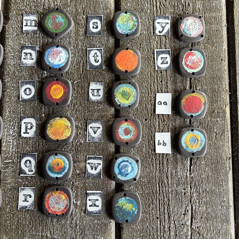 May include: A collection of 26 ceramic tiles with colourful designs and letters from the alphabet. Each tile has a different colour and pattern, and the letters are printed in black.