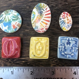 May include: Six ceramic tiles with colorful designs. Two oval tiles with abstract patterns in shades of blue, green, yellow, orange, and red. Four square tiles with letters O, U, and V in raised relief. The O tile is red, the U tile is yellow, and the V tile is blue.