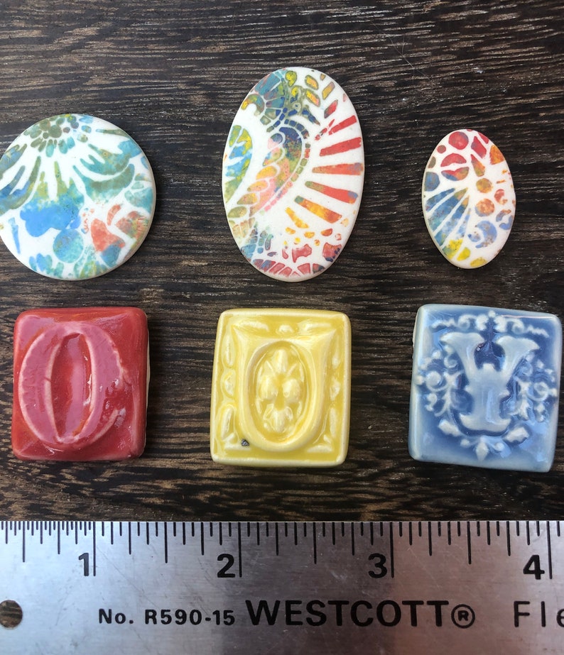 May include: Six ceramic tiles with colorful designs. Two oval tiles with abstract patterns in shades of blue, green, yellow, orange, and red. Four square tiles with letters O, U, and V in raised relief. The O tile is red, the U tile is yellow, and the V tile is blue.