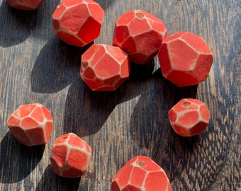 May include: A selection of red, geometric beads with white detailing and a hole for threading. The beads are displayed on a dark wooden surface, with shadows from sunlight. The beads have a faceted design.