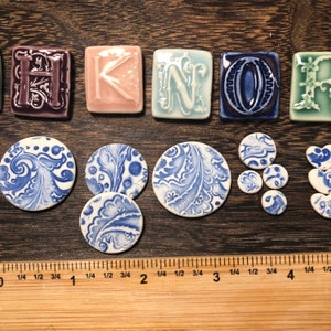 May include: A selection of ceramic tiles and decorative elements. The tiles spell "HOME" in shades of purple, pink, blue, and green. There are also round and heart-shaped pieces with blue and white floral designs. A ruler is visible, with measurements in inches.