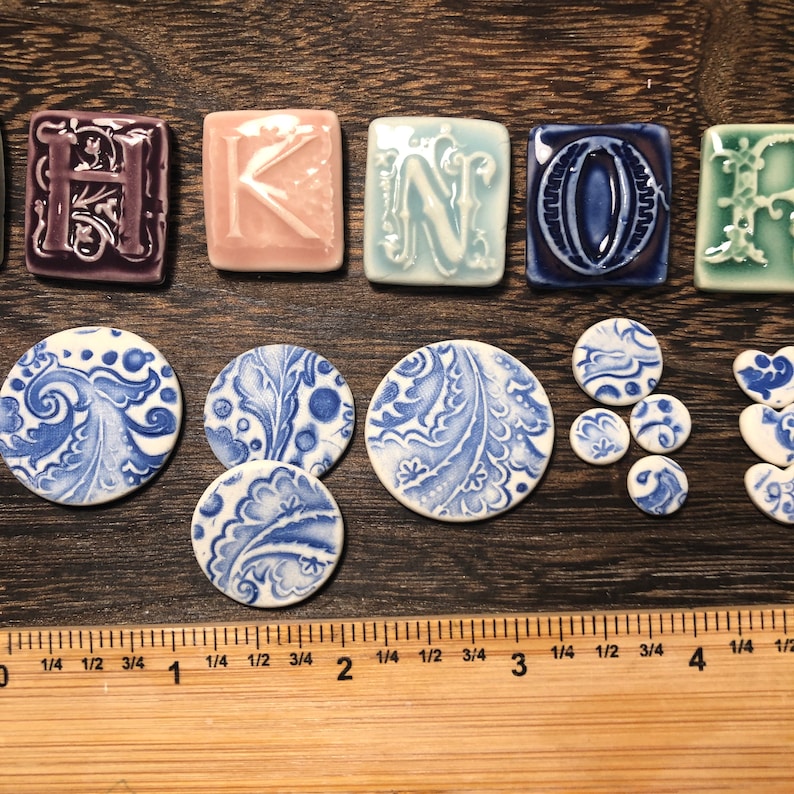 May include: A selection of ceramic tiles and decorative elements. The tiles spell "HOME" in shades of purple, pink, blue, and green. There are also round and heart-shaped pieces with blue and white floral designs. A ruler is visible, with measurements in inches.