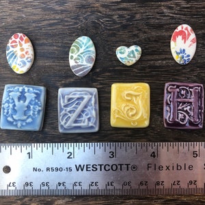 May include: A set of nine ceramic tiles featuring different designs. The tiles are in various colors, including blue, purple, yellow, and white. The tiles are arranged in a row on a wooden surface. The tiles are numbered 1 through 5. The tiles are all approximately the same size and shape. The tiles are all handmade and unique.