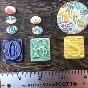May include: A set of ceramic tiles with colorful designs. The tiles are in the shape of circles, ovals, and squares. The tiles feature a variety of patterns, including floral, geometric, and abstract designs. The tiles are arranged on a wooden surface with a ruler for scale. The ruler is labeled "WESTCOTT" and has markings in inches.